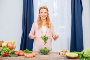Woman in a pink shirt preparing spinach salad with various fresh vegetables on a kitchen counter.