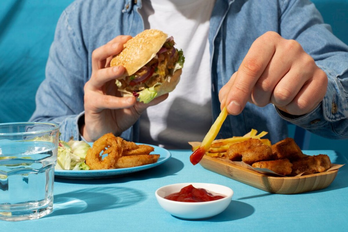 Person dipping a french fry in ketchup with a burger, fried fish, and glass of water on a blue table.