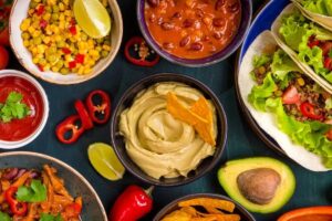 Assorted Mexican dishes including tacos, guacamole, corn salad, and chili beans on a dark table.