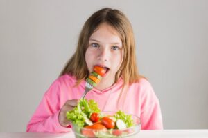 Young girl in pink shirt eating a salad with a fork, looking at the camera.