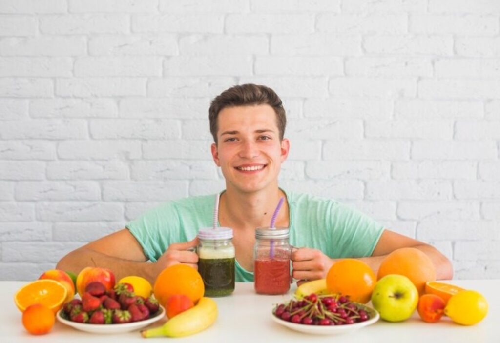 Young man smiling at a table with assorted fruits and two smoothies.