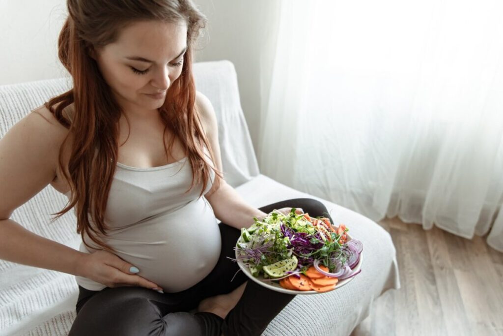 Pregnant woman sitting on a couch, looking at a colorful salad she is holding.