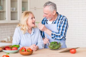 Senior man feeding a leafy green to a woman in a kitchen filled with fresh vegetables.