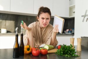 Young woman looking confused with vegetables and a recipe book in a modern kitchen.