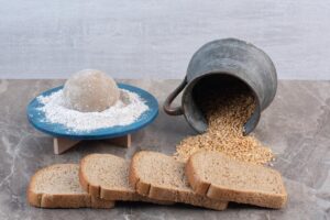 Bread dough on a blue plate, overturned bucket of grains, and sliced bread on a marble surface.