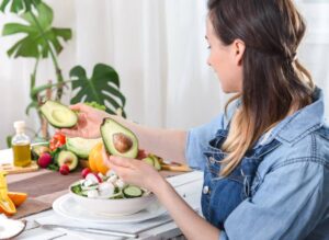 Woman in denim shirt preparing a salad with fresh vegetables and avocado in a bright kitchen.