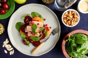 A healthy meal of sliced beets, oranges, spinach, and turkey slices garnished with pomegranate on a white plate.
