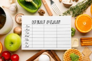 Blank meal plan surrounded by fresh fruits, vegetables, and various kitchen tools on a white surface.