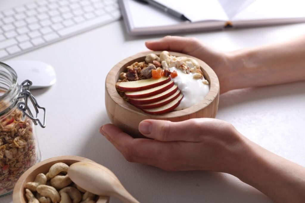Person holding a wooden bowl with yogurt, granola, nuts, and apple slices, at a desk with computer accessories.