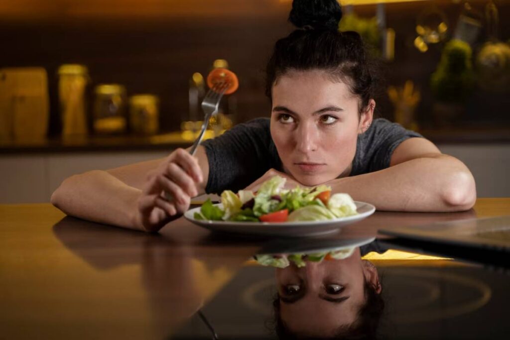 Woman thoughtfully looking at salad with its reflection on table, in a dimly lit room.