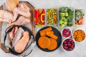 Assorted raw meats and breaded cutlets with containers of colorful vegetables and frozen berries, displayed for meal preparation.