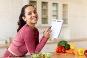 Smiling woman in pink athletic wear writing in a notebook labeled Diet plan with fresh vegetables and salad on table.
