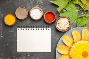 Various spices in wooden bowls, shredded chicken, an open notebook, and a dish with corn chips and dip, on a dark table.