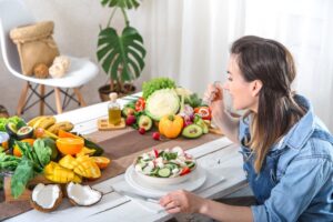 Woman in denim shirt eating fresh salad at a table filled with various fruits and vegetables.